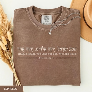 May include: An espresso-colored crew neck t-shirt featuring white text that reads: Hear, O Israel: The Lord our God, the Lord is one, Deuteronomy 6:4, along with the Hebrew translation of the verse. A silver star pendant necklace rests on the collar.