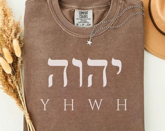 Puede incluir: Una camiseta de cuello redondo color espresso que presenta los caracteres hebreos para YHWH en un tono crema claro, con las letras inglesas Y H W H impresas debajo en una fuente y color a juego. Un collar con dije de estrella plateado descansa sobre el cuello.