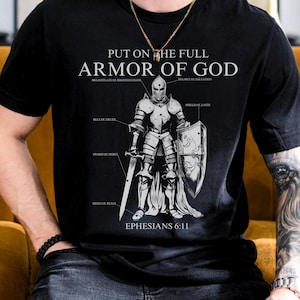May include: A black short-sleeve t-shirt featuring a white graphic of a knight in armor with labels pointing to parts labeled Breastplate of Righteousness, Helmet of Salvation, Belt of Truth, Sword of Spirit, and Shield of Faith, with text Put on the Full Armor of God and Ephesians 6:11.