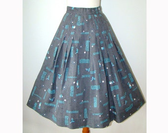 May include: A high-waisted, pleated midi skirt in a dark grey fabric featuring a repeating pattern of small light blue figures, white circular dots, and rectangular blocks of light blue text distributed across the entire surface of the garment.