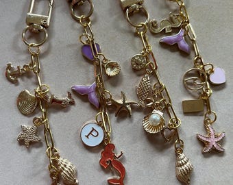 May include: Four gold-colored keychains with various charms. Charms include seashells, starfish, a sailboat, a mermaid, and a heart. Some charms are accented with pink, purple, and red enamel. Each keychain has a lobster clasp.
