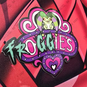 May include: A colorful sticker featuring a cartoon frog with the text "Froggie's". The sticker has a purple and pink heart design with a green and purple frog character. The background is a red and black geometric pattern.