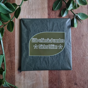 Könnte beinhalten: Ein dunkelgrünes Paket mit einem Aufkleber mit der Aufschrift "Little whimsical wonders Sticker Edition" in weißem Text. Grüne, belaubte Ranken umrahmen das Paket, vor einem Holzgrund.
