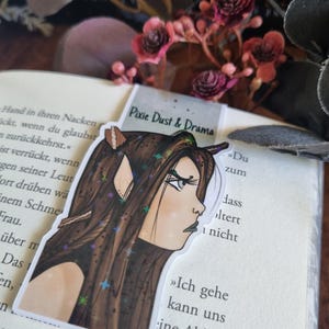 Könnte beinhalten: Ein Aufkleber mit dem Profil einer Frau mit langen braunen Haaren, spitzen Ohren und Sternen. Der Aufkleber trägt den Text "Pixie Dust & Drama" und befindet sich auf einem offenen Buch mit deutschem Text.