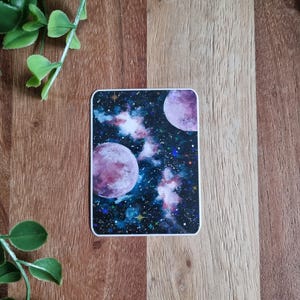 Könnte beinhalten: Ein rechteckiger Aufkleber mit einem kosmischen Design. Er zeigt zwei große, violette Planeten, umgeben von einem lebendigen Nebel aus Blau-, Rosa- und Lilatönen, mit Sternen und Himmelselementen.