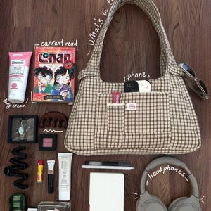 May include: A beige and brown plaid shoulder bag surrounded by various items including a Detective Conan manga, a Balea body lotion tube, a white notebook, grey over-ear headphones, a perfume bottle, sunglasses, and assorted small cosmetic items.