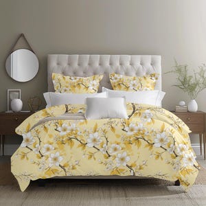 May include: A bed dressed with a yellow floral duvet cover and pillowcases. The design shows white flowers and gold leaves on a yellow backdrop. White pillows and a buttoned headboard complete the bedding set.