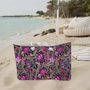 Peut inclure: Un grand sac fourre-tout avec un motif floral dans les tons de violet, rose, vert et marron. Le sac a des poignées en corde blanche et est posé sur une plage de sable. L'arrière-plan montre une scène de plage avec l'océan et une cabane.