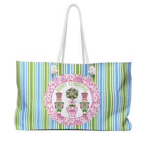 Può includere: Borsa tote di grandi dimensioni con strisce verticali blu e verdi. La borsa presenta un design rotondo con due schiaccianoci, una topiaria e le parole "MERRY CHRISTMAS" in una cornice floreale rosa. I manici sono bianchi.