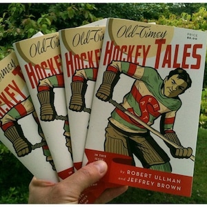 May include: A stack of four comic books with a hockey theme. The cover of the top book features a hockey player in a green and white jersey with a red and white stripe. The title of the book is "Old-Timey Hockey Tales".