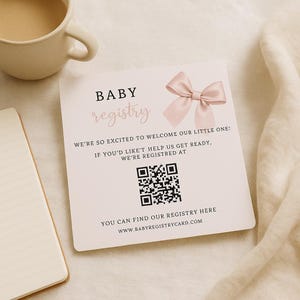 Könnte beinhalten: Eine quadratische Baby-Registrierungskarte mit einer rosa Schleife und den Worten "BABY registry". Die Karte enthält den Text "WE'RE SO EXCITED TO WELCOME OUR LITTLE ONE!" und einen QR-Code. Eine Tasse Kaffee und ein Notizblock sind im Hintergrund.