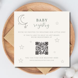 May include: A baby registry card with the text "BABY registry" and the phrase "WE'RE SO EXCITED TO WELCOME OUR LITTLE ONE!". The card features a QR code and star and moon illustrations. The card is on a wooden plate.