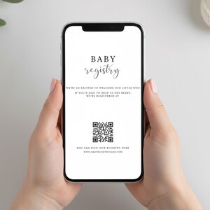 May include: A smartphone displays a baby registry announcement. The screen features the words "BABY registry" in elegant script, along with a QR code and website address. The phone is held against a white background, with a sprig of greenery visible.