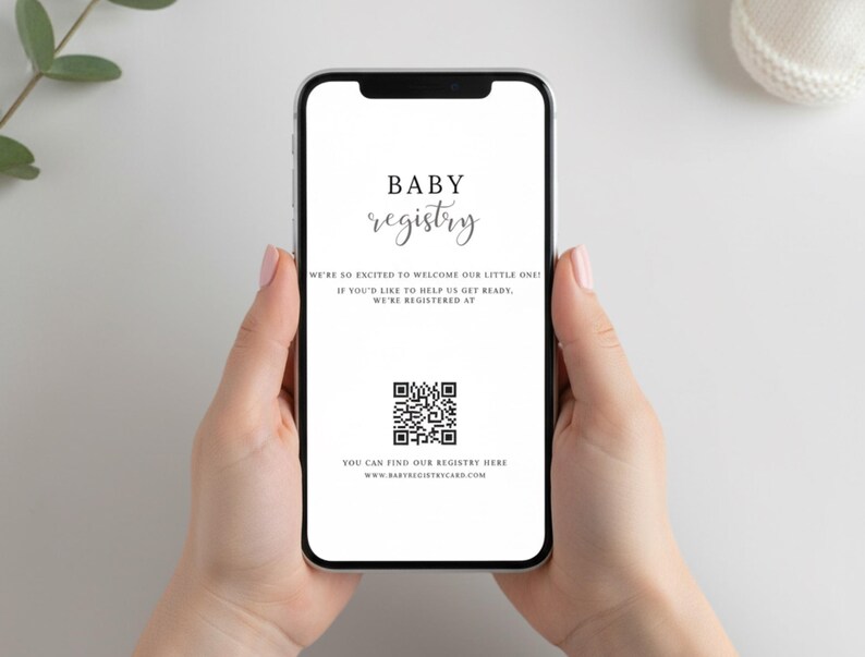 May include: A smartphone displays a baby registry announcement. The screen features the words "BABY registry" in elegant script, along with a QR code and website address. The phone is held against a white background, with a sprig of greenery visible.