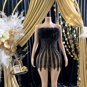 Pode incluir: Um vestido preto de lantejoulas com uma borda de penas no decote. O vestido é transparente e tem uma silhueta ajustada. O vestido é exibido em um manequim contra um fundo de tecido dourado e preto.