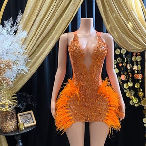 Puede incluir: Vestido mini naranja y transparente con cuentas, escote pronunciado y ribete de plumas. El vestido está en un maniquí.