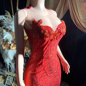 May include: A red, floor-length gown with intricate beadwork and a sweetheart neckline. The bodice features a sheer overlay and detailed embellishments. The dress is displayed on a mannequin, showcasing its form-fitting silhouette and flared hemline, perfect for formal events.