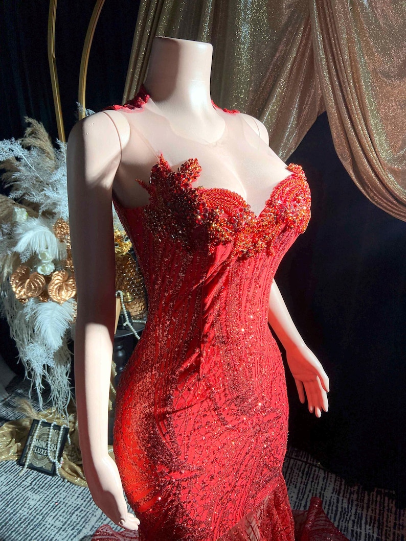 May include: A red, floor-length gown with intricate beadwork and a sweetheart neckline. The bodice features a sheer overlay and detailed embellishments. The dress is displayed on a mannequin, showcasing its form-fitting silhouette and flared hemline, perfect for formal events.