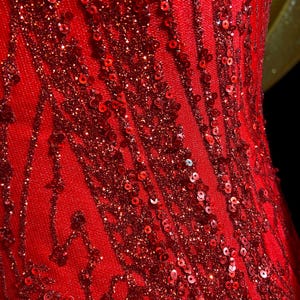 May include: Close-up of a vibrant red dress with intricate sequin and glitter detailing. The fabric features vertical and diagonal patterns, creating a textured and shimmering effect. The dress appears to be made of a soft, flowing material.