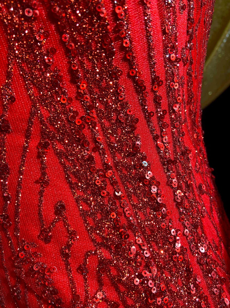 May include: Close-up of a vibrant red dress with intricate sequin and glitter detailing. The fabric features vertical and diagonal patterns, creating a textured and shimmering effect. The dress appears to be made of a soft, flowing material.