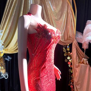 May include: A red sequined evening gown on a mannequin. The dress features a sheer neckline and intricate beadwork on the bodice. The gown has a fitted silhouette that flares out at the bottom. The background includes gold drapery and decorative elements.
