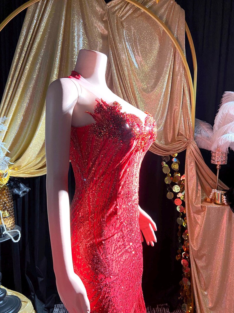May include: A red sequined evening gown on a mannequin. The dress features a sheer neckline and intricate beadwork on the bodice. The gown has a fitted silhouette that flares out at the bottom. The background includes gold drapery and decorative elements.
