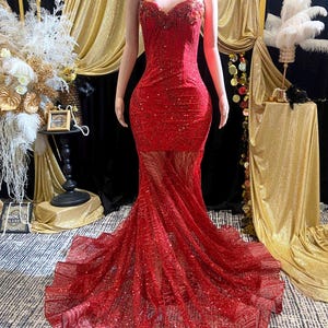 May include: A red sequined formal gown on a mannequin. The dress features a sweetheart neckline, a fitted bodice, and a flowing mermaid skirt. The gown is embellished with sparkling sequins and a sheer overlay, creating a glamorous look.