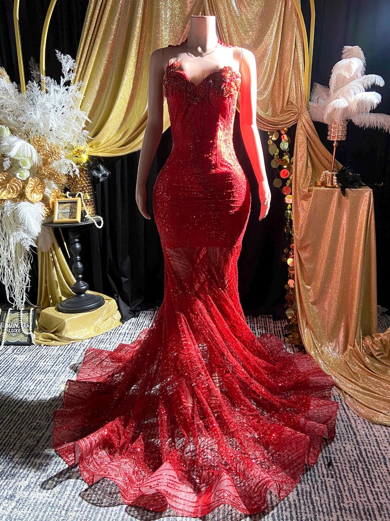 May include: A red, floor-length gown on a mannequin. The dress features a sweetheart neckline, fitted bodice, and a flowing, sheer skirt. The gown is embellished with sequins and intricate detailing, creating a glamorous look. The background includes gold and black drapery.
