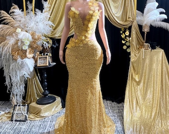 May include: A gold sequined formal gown on a mannequin. The dress features a one-shoulder design with floral embellishments and a sheer panel. The gown has a mermaid silhouette and is floor-length.
