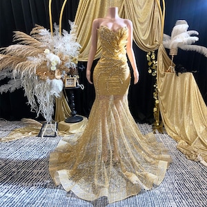 May include: A gold, floor-length formal gown with a sweetheart neckline and a mermaid silhouette. The bodice is embellished with sequins and a feathered design. The skirt features a sheer, flowing overlay, creating a glamorous look.