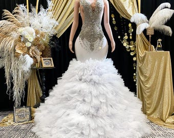 May include: A white mermaid-style gown with a heavily embellished bodice and a ruffled skirt. The bodice is adorned with silver-coloured beading. The dress is displayed on a mannequin against a backdrop of gold and black.