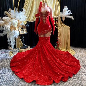 Puede incluir: Un vestido rojo con lentejuelas, corpiño transparente y una larga cola fluida. El vestido incluye guantes largos y un tocado a juego. El vestido se exhibe en un maniquí, mostrando su intrincado diseño y detalles brillantes. Es un vestido de fiesta.