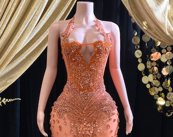 May include: A sheer orange mini dress with a halter neckline and intricate beading throughout. The dress is fitted and has a form-fitting silhouette.