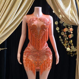 May include: A short, sleeveless dress with a sheer fabric base. The dress is covered in orange sequins and beads, with fringe detailing around the neckline and hem.