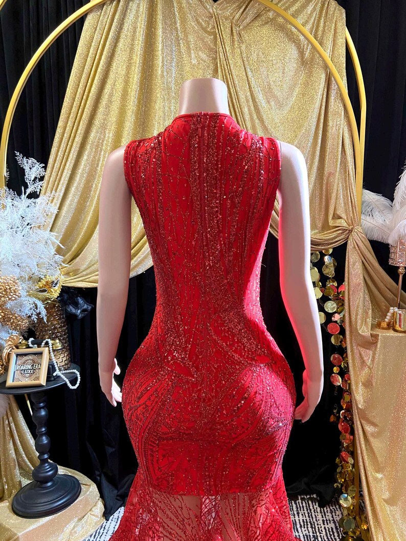 May include: A red, sleeveless, floor-length gown with intricate sequin detailing. The dress features a high neckline and a mermaid silhouette. The back of the dress is visible, showcasing the design. The dress is displayed on a mannequin.