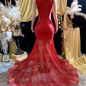 May include: A red, floor-length gown with a high neckline and a sheer, flowing skirt. The dress is covered in sequins and sparkles, creating a glamorous look. The back of the dress is visible, showcasing the intricate design and fit.