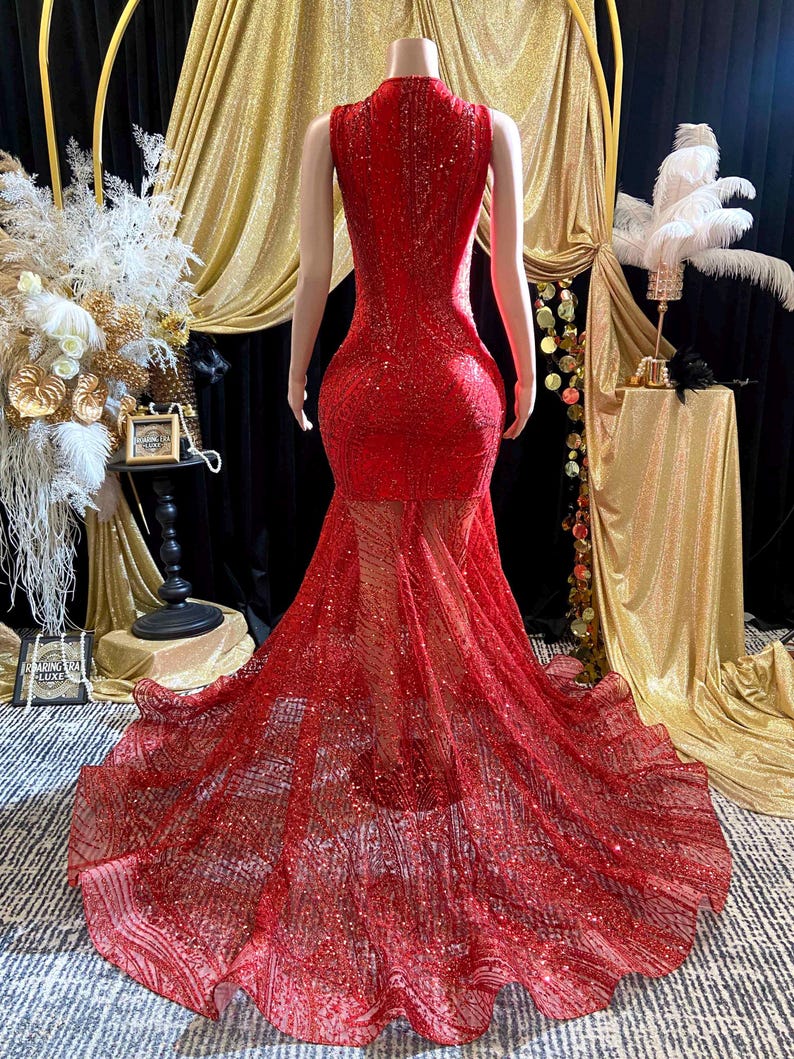 May include: A red, floor-length gown with a high neckline and a sheer, flowing skirt. The dress is covered in sequins and sparkles, creating a glamorous look. The back of the dress is visible, showcasing the intricate design and fit.