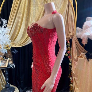 May include: A vibrant red formal gown with intricate beadwork and a fitted silhouette. The dress features a halter neckline and a detailed bodice, perfect for special occasions. The gown is displayed on a mannequin, showcasing its elegant design and texture.