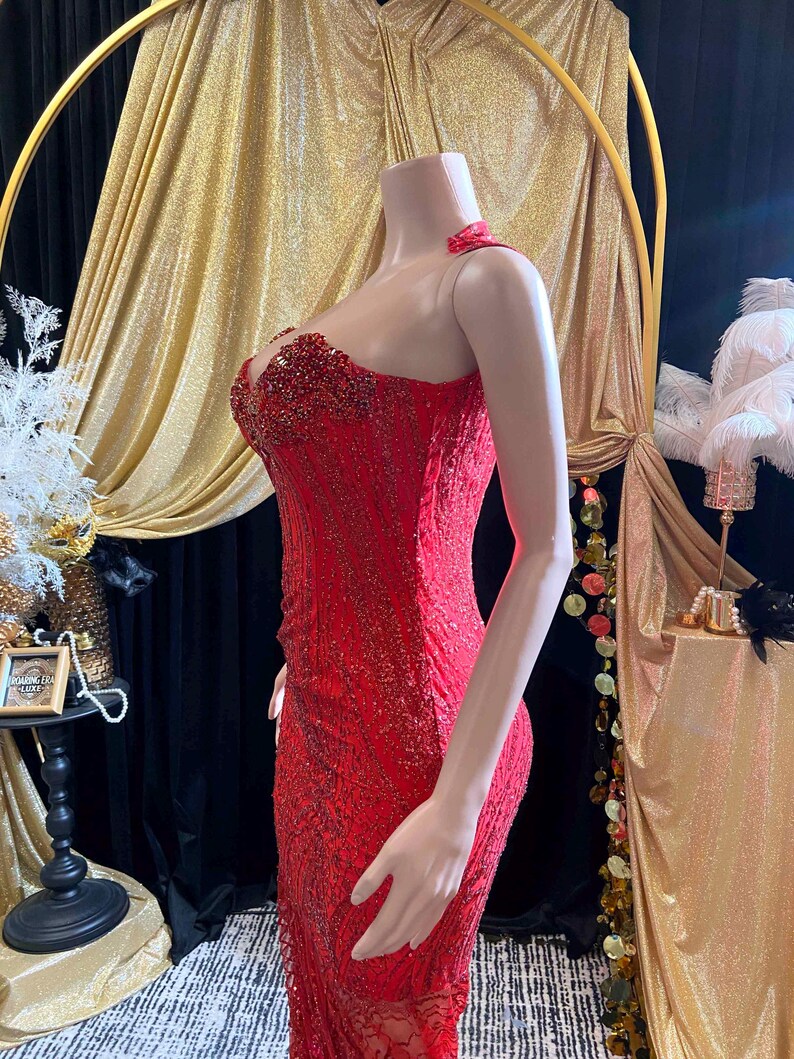 May include: A vibrant red formal gown with intricate beadwork and a fitted silhouette. The dress features a halter neckline and a detailed bodice, perfect for special occasions. The gown is displayed on a mannequin, showcasing its elegant design and texture.