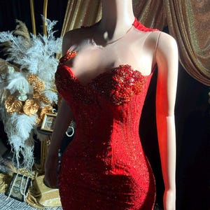 May include: A red, beaded evening gown on a headless mannequin. The dress features a sweetheart neckline, intricate beadwork, and a sheer neckline. The gown is form-fitting with a mermaid silhouette, and is displayed in front of a gold backdrop.