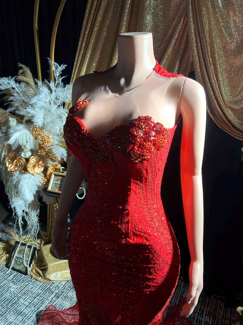 May include: A red, beaded evening gown on a headless mannequin. The dress features a sweetheart neckline, intricate beadwork, and a sheer neckline. The gown is form-fitting with a mermaid silhouette, and is displayed in front of a gold backdrop.