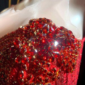 May include: Close-up of a red, embellished bodice. The bodice is covered in red sequins and large, faceted red gems. Gold-colored sequins are interspersed throughout the design. The garment appears to be part of a formal dress or gown.