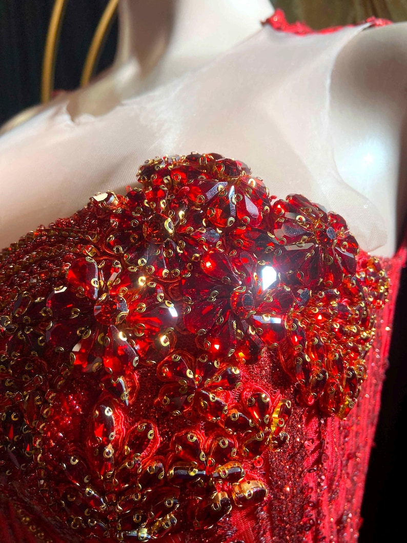 May include: Close-up of a red, embellished bodice. The bodice is covered in red sequins and large, faceted red gems. Gold-colored sequins are interspersed throughout the design. The garment appears to be part of a formal dress or gown.