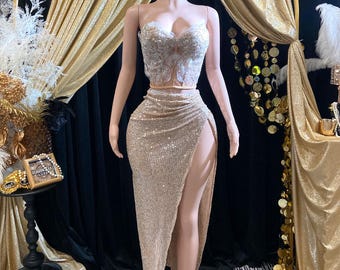 May include: A gold sequined skirt with a high slit and a white lace crop top. The skirt and top are worn by a mannequin against a backdrop of gold fabric.
