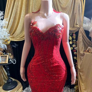 May include: A vibrant red, floor-length gown with intricate sequin and bead detailing. The dress features a sweetheart neckline, a sheer upper bodice, and a mermaid silhouette. The gown is displayed on a mannequin.