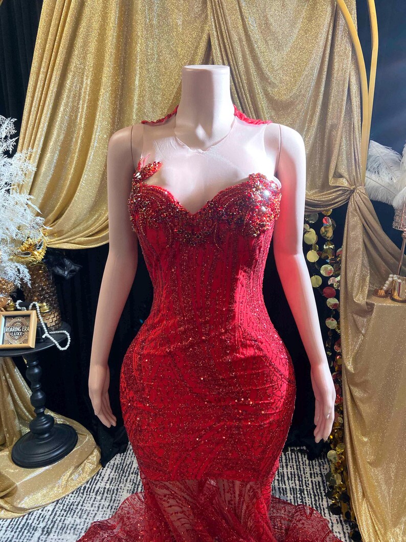 May include: A vibrant red, floor-length gown with intricate sequin and bead detailing. The dress features a sweetheart neckline, a sheer upper bodice, and a mermaid silhouette. The gown is displayed on a mannequin.