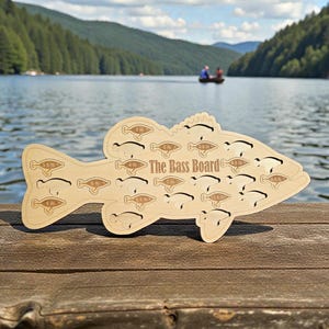 May include: A wooden fish-shaped board with the text "The Bass Board" and several fish silhouettes. Each silhouette has a weight measurement in pounds. The board is light brown and sits on a wooden surface with a lake and mountains in the background.