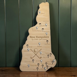 May include: A wooden map of New Hampshire with engraved text reading "New Hampshire Top Public Golf Courses." The map features golf course pin markers and crossed golf clubs with a golf ball. The map is made of light-colored wood.