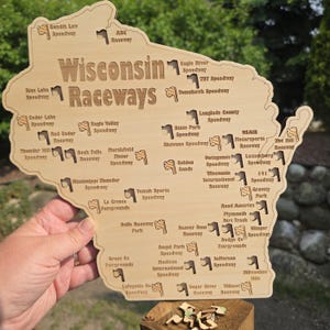 May include: A wooden map of Wisconsin with the words "Wisconsin Raceways" engraved. The map features the names and locations of various speedways across the state, marked with small checkered flag icons. The map is held by a hand.