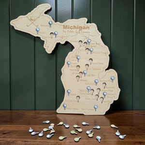 May include: Wooden map of Michigan with golf course markers. The map is engraved with the text "Michigan Top Public Golf Courses" and features small golf ball markers. A golf club and ball are at the top.